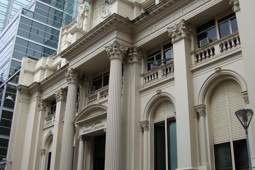 Fachada de piedra clara con columnas, balcones y faroles de un edificio histórico, con una torre vidriada detrás y la bandera argentina en lo alto.