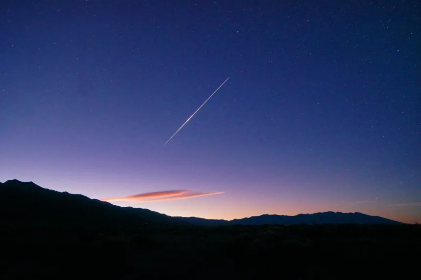 Una raya de luz blanca cruza en diagonal un cielo azul oscuro con estrellas, sobre una sierra en silueta y un horizonte anaranjado.