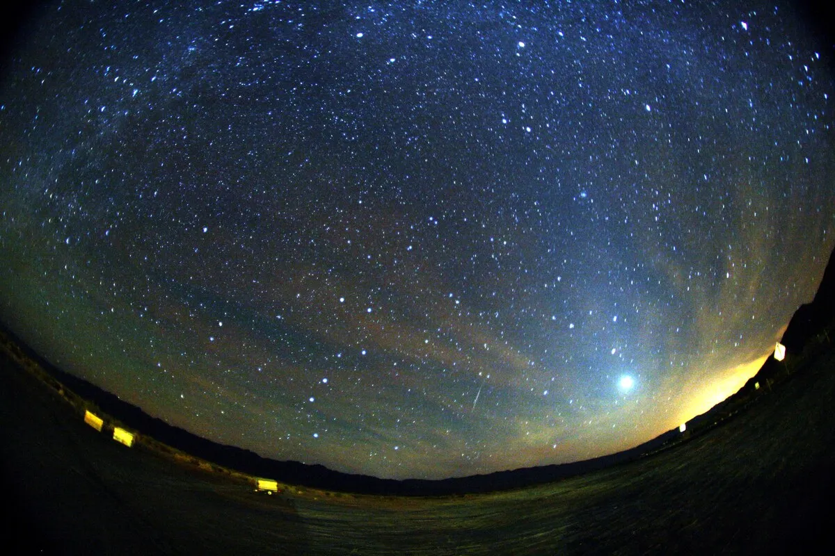 Cielo estrellado visto con lente de ojo de pez, con la Vía Láctea, un meteoro verde y rojo y un planeta brillante sobre el horizonte.
