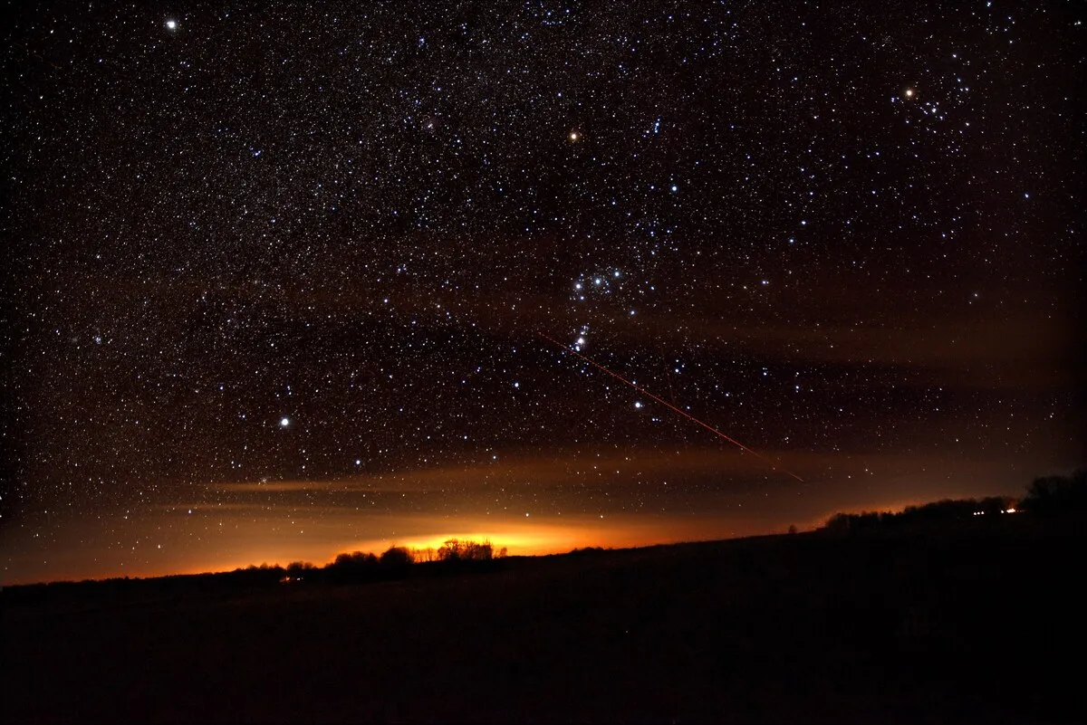 Cielo nocturno con nubes finas y un resplandor anaranjado en el horizonte; en el centro, las tres estrellas alineadas del cinturón de Orión.