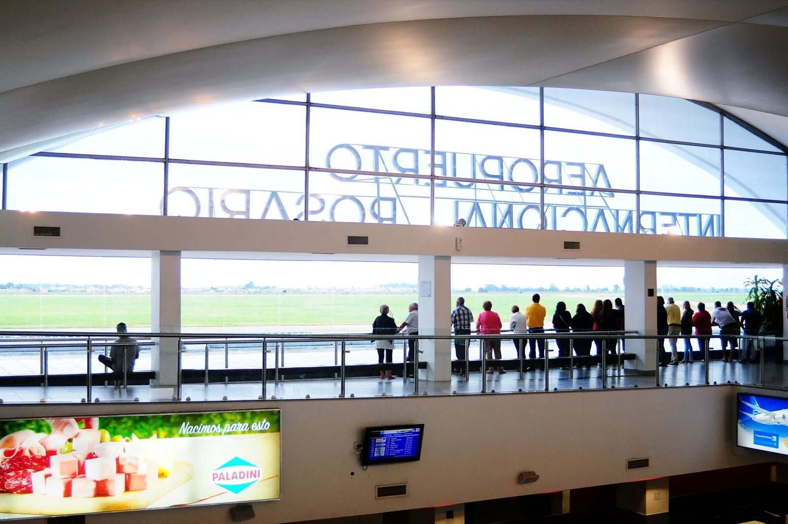 Hall de un aeropuerto con un ventanal hacia la pista, gente asomada a una baranda y letras grandes que dicen «Aeropuerto Internacional Rosario».