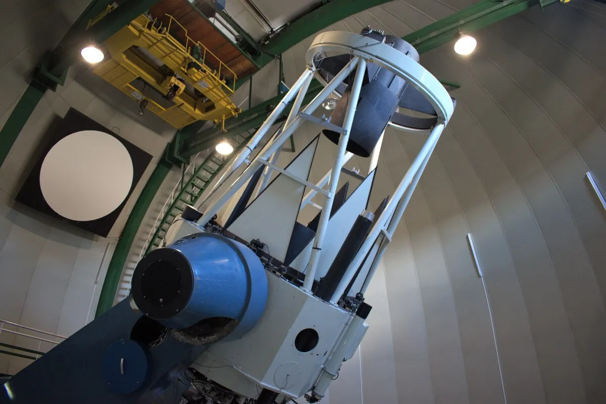 Telescopio grande de estructura blanca y azul, visto desde abajo dentro de una cúpula.