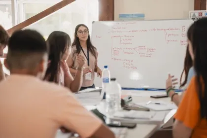 Una docente da clase frente a una pizarra y varios estudiantes la escuchan.