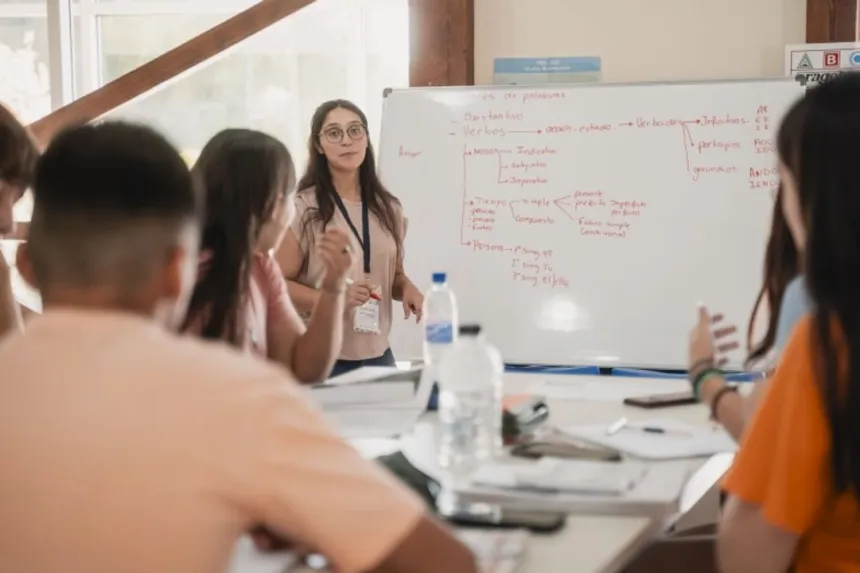 Una docente da clase frente a una pizarra y varios estudiantes la escuchan.