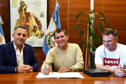Tres hombres firman un convenio en una oficina con banderas argentinas y de San Juan.