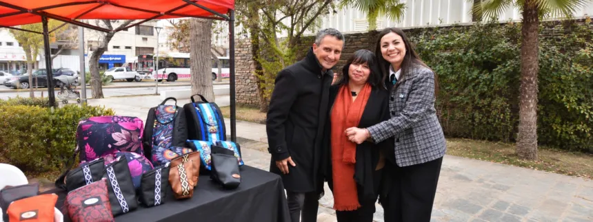 Tres adultos junto a una mesa con bolsos y productos artesanales.