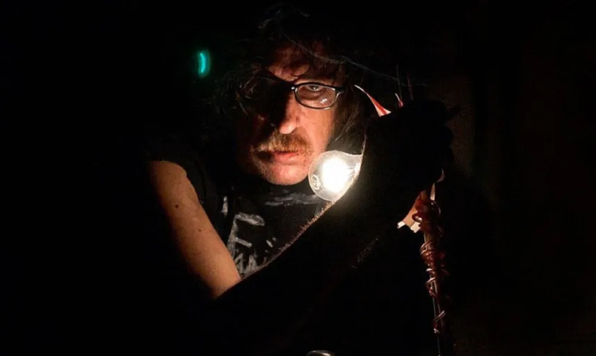 Charly García, con gafas, junto a un micrófono en una escena oscura.