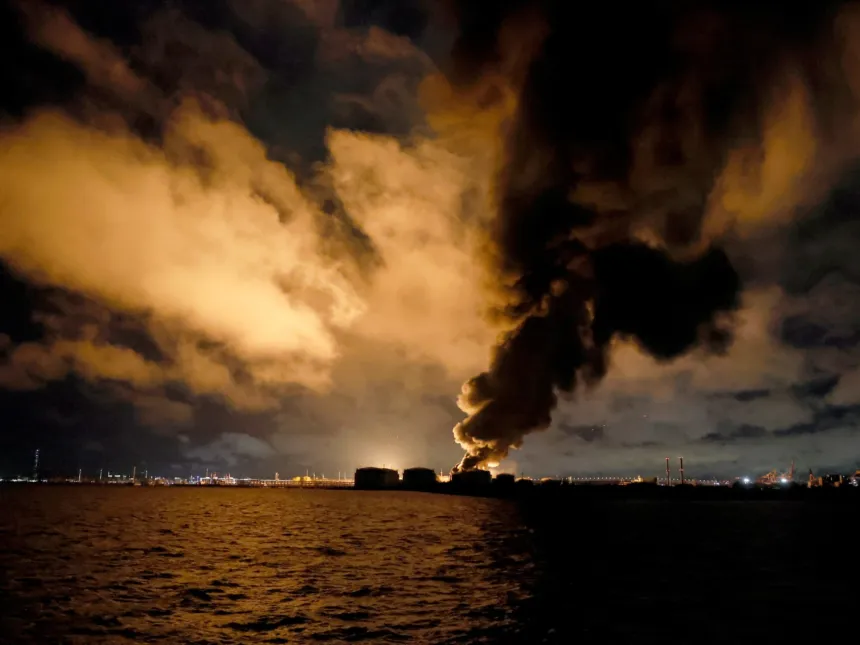 Una instalación industrial junto al agua arde de noche y despide una gran columna de humo.