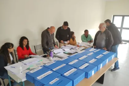 Personas adultas organizan una votación junto a urnas azules identificadas con nombres de canales.