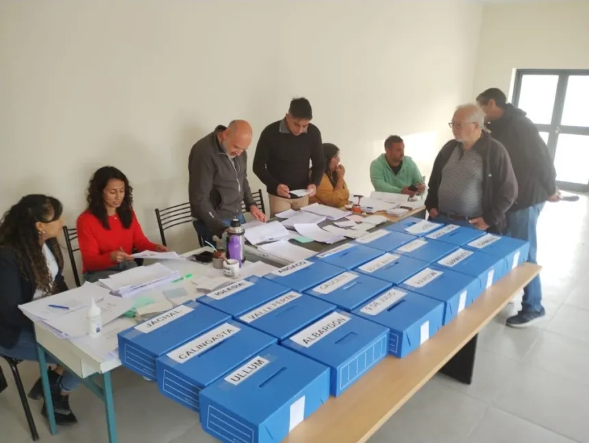 Personas adultas organizan una votación junto a urnas azules identificadas con nombres de canales.