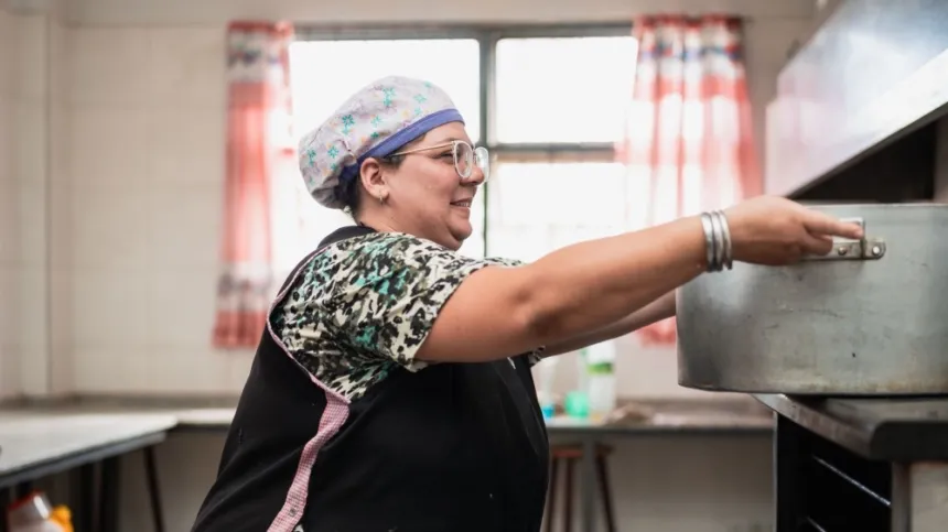 Una trabajadora con uniforme cocina junto a una olla grande.