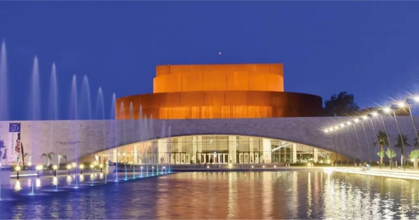 Teatro iluminado de noche, con fuentes y agua en primer plano.