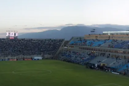 Público en las tribunas del estadio del Bicentenario junto a una cancha de fútbol al atardecer.