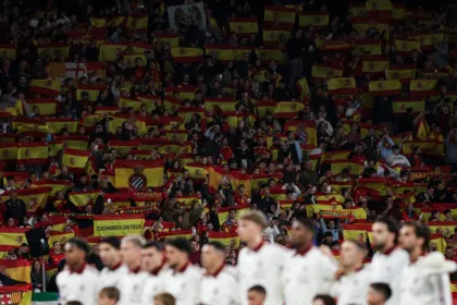 Hinchas con banderas de España en una tribuna, detrás de jugadores desenfocados.