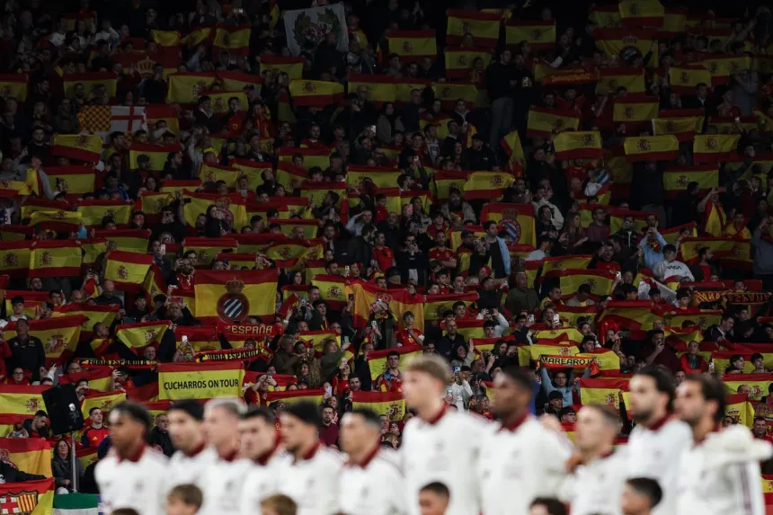 Hinchas con banderas de España en una tribuna, detrás de jugadores desenfocados.