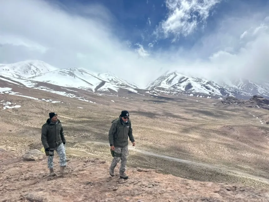 Dos gendarmes recorren una ladera rocosa en la alta montaña, con cumbres nevadas al fondo.
