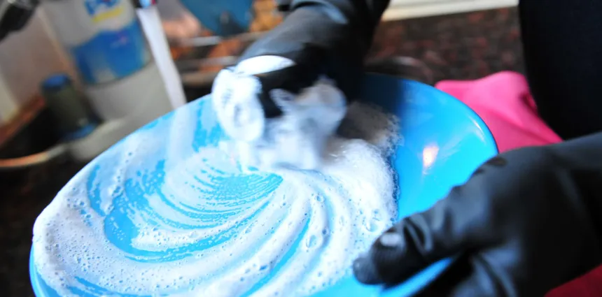 Manos con guantes limpian un objeto en un recipiente azul con agua jabonosa.