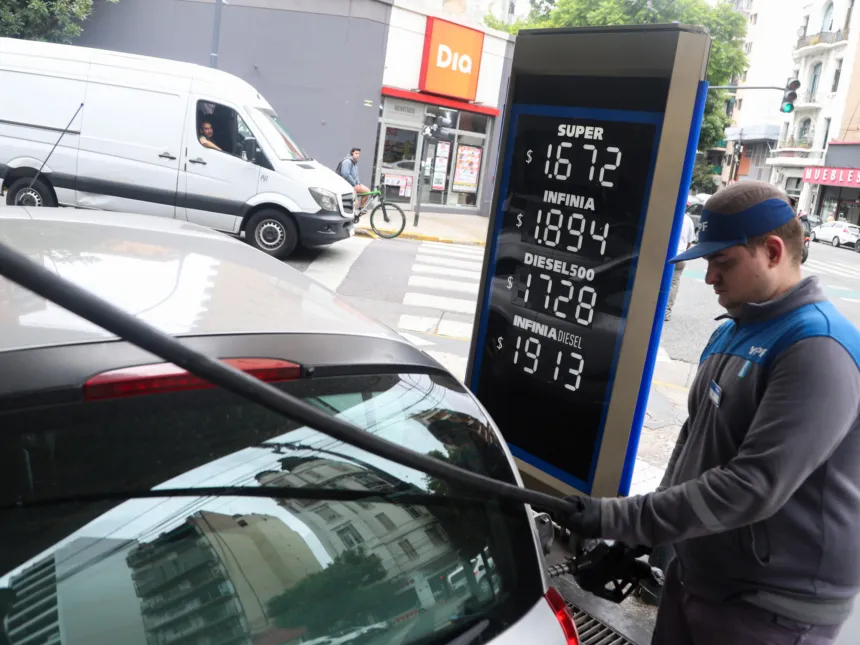 Un empleado carga combustible a un auto junto al cartel de precios de la estación.