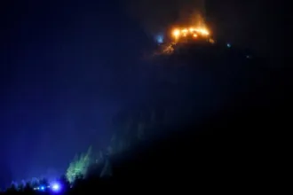Un edificio en una montaña arde de noche; luces de emergencia iluminan la ladera.