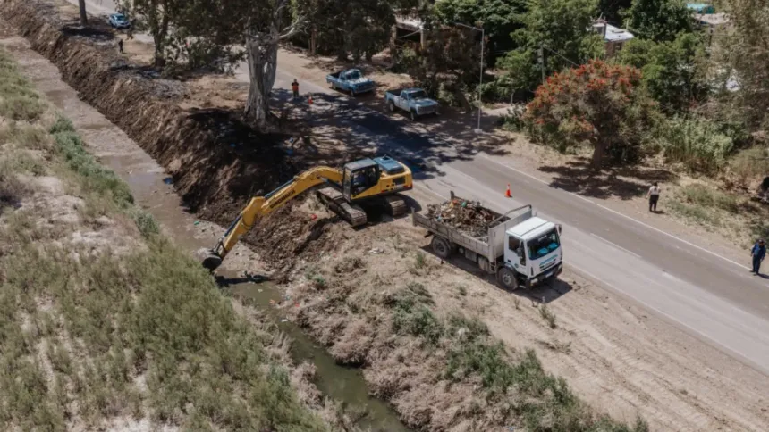 Una excavadora limpia un desagüe junto a una ruta y un camión carga los sedimentos.