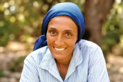 Mujer sonriente con pañuelo azul en la cabeza, retratada al aire libre.