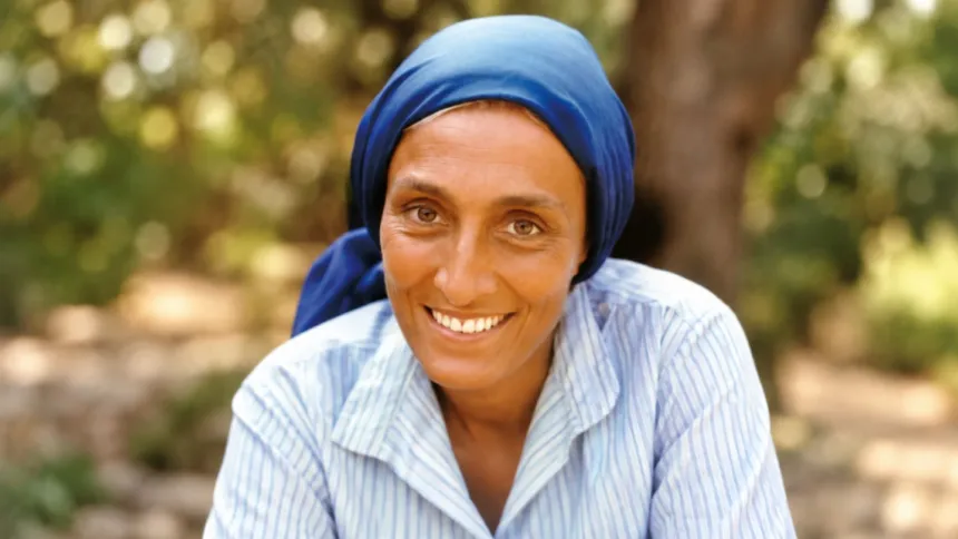 Mujer sonriente con pañuelo azul en la cabeza, retratada al aire libre.