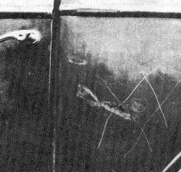 Primer plano en blanco y negro de la puerta de un auto oscuro, con la manija arriba a la izquierda y varias rayas finas y curvas grabadas en la chapa.