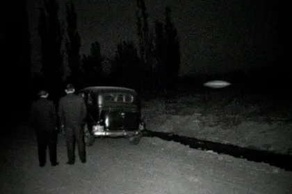Recreación en blanco y negro con aspecto de foto de 1968: dos hombres de traje, de espaldas, junto a un auto antiguo en una calle de tierra con álamos y una acequia, de noche; sobre un baldío flota un objeto ovalado borroso con un brillo tenue.