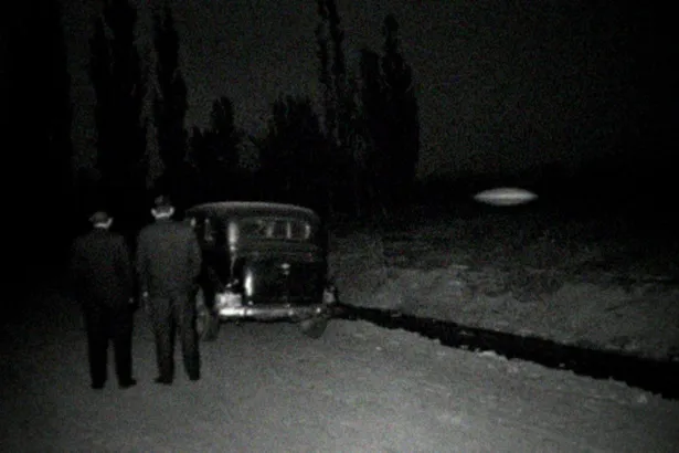 Recreación en blanco y negro con aspecto de foto de 1968: dos hombres de traje, de espaldas, junto a un auto antiguo en una calle de tierra con álamos y una acequia, de noche; sobre un baldío flota un objeto ovalado borroso con un brillo tenue.