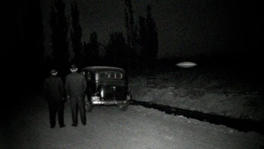 Recreación en blanco y negro con aspecto de foto de 1968: dos hombres de traje, de espaldas, junto a un auto antiguo en una calle de tierra con álamos y una acequia, de noche; sobre un baldío flota un objeto ovalado borroso con un brillo tenue.