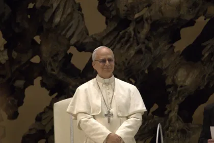El papa León XIV, con sotana blanca y cruz pectoral, sonríe de pie frente a una escultura de bronce en el Vaticano.