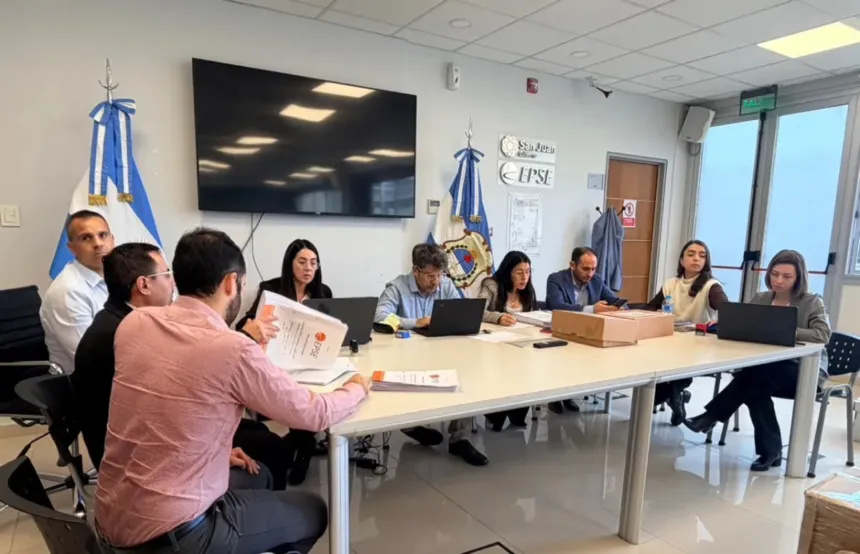 Personas revisan documentos y computadoras durante una reunión en una sala con banderas.