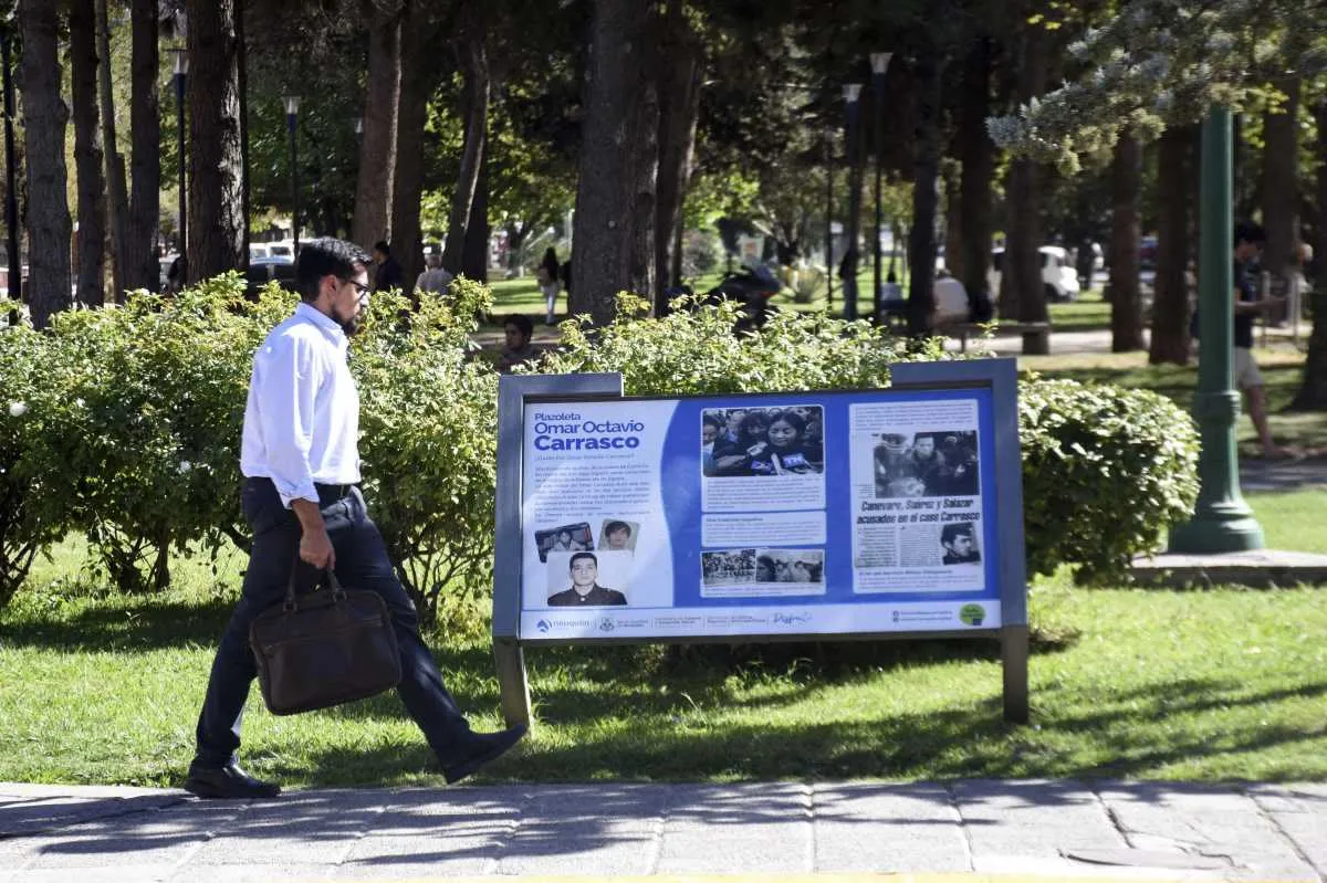Un hombre camina frente a un panel con fotos y textos sobre Omar Carrasco en una plaza arbolada.
