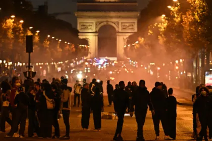 Multitud reunida entre humo frente al Arco del Triunfo de noche.