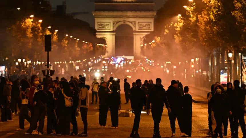 Multitud reunida entre humo frente al Arco del Triunfo de noche.