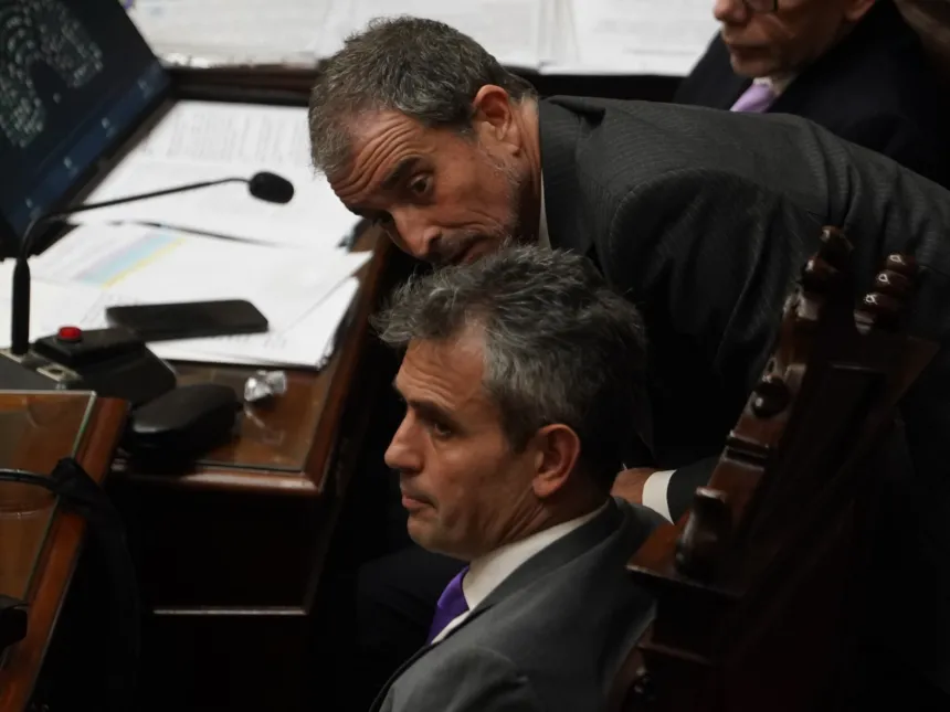 Dos hombres conversan en sus bancas durante una sesión legislativa.