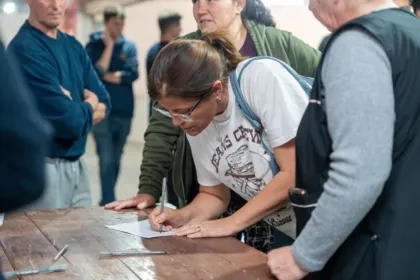 Una mujer firma un documento en una mesa, rodeada de otras personas.