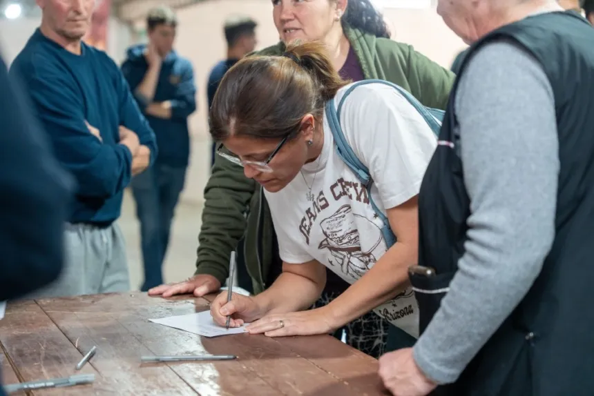 Una mujer firma un documento en una mesa, rodeada de otras personas.
