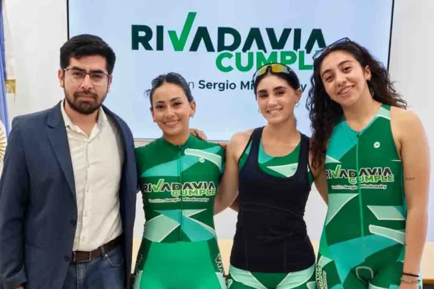 Tres deportistas con indumentaria de Rivadavia posan junto a un hombre frente a un cartel municipal.