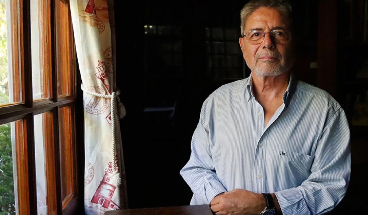 Hombre mayor de anteojos y camisa celeste, sentado junto a una ventana.