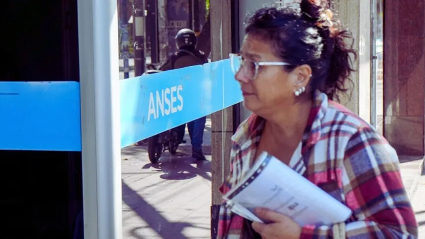 Una mujer adulta con papeles frente a un cartel de ANSES.