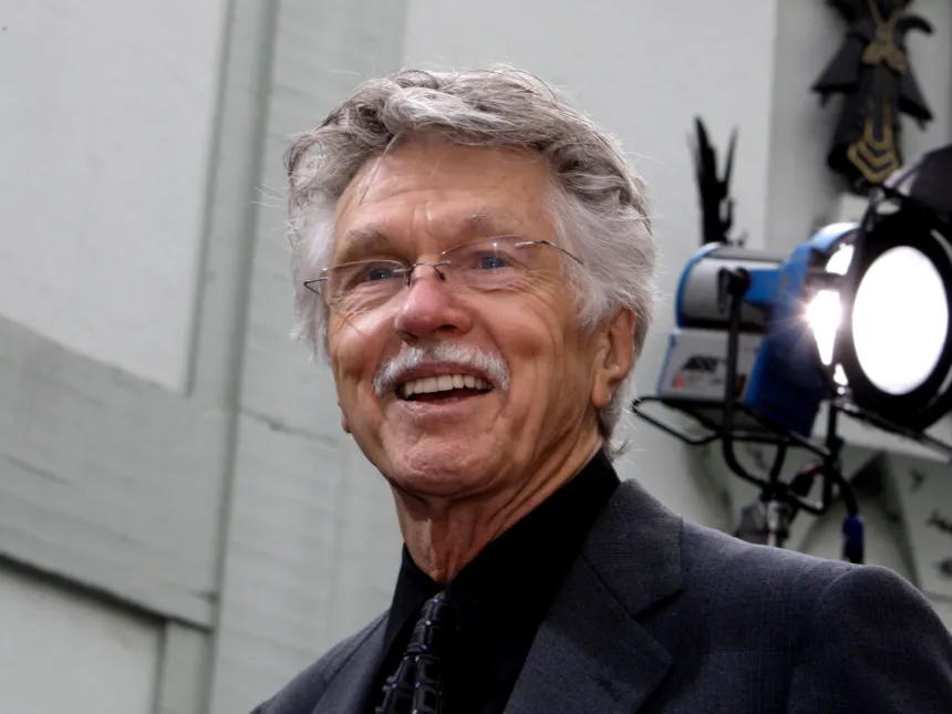 Tom Skerritt sonríe vestido de traje junto a un equipo de iluminación.