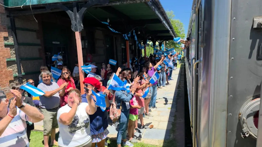 Personas saludan con banderas argentinas a un tren desde el andén.